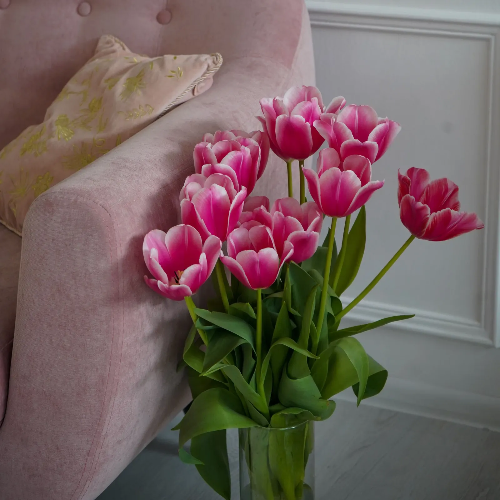 Strauß pinker Tulpen in einer Glasvase neben einem rosa Samtsessel vor weißer Wand